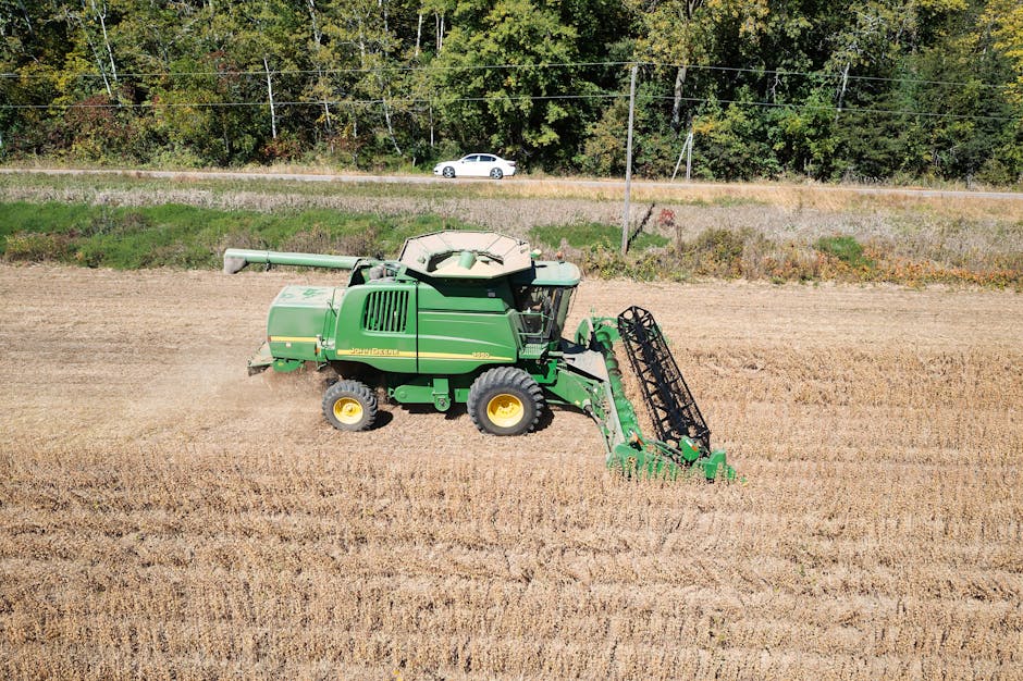 A green John Deere combine harvester is actively working across a large, flat agricultural field with freshly tilled soil, creating evenly spaced lines in the earth. The machinery’s large black tire