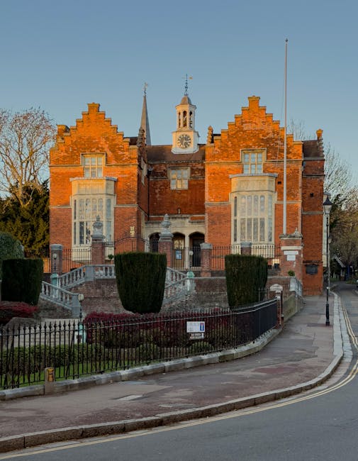 A historic red brick building with ornate architectural details, featuring stepped gables, tall windows, and a central clock tower with a small spire, situated in a well-maintained garden. The garden 