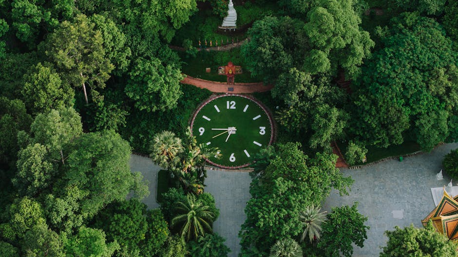 A historic red brick building with ornate architectural details, featuring stepped gables, tall windows, and a central clock tower with a small spire, situated in a well-maintained garden. The garden 